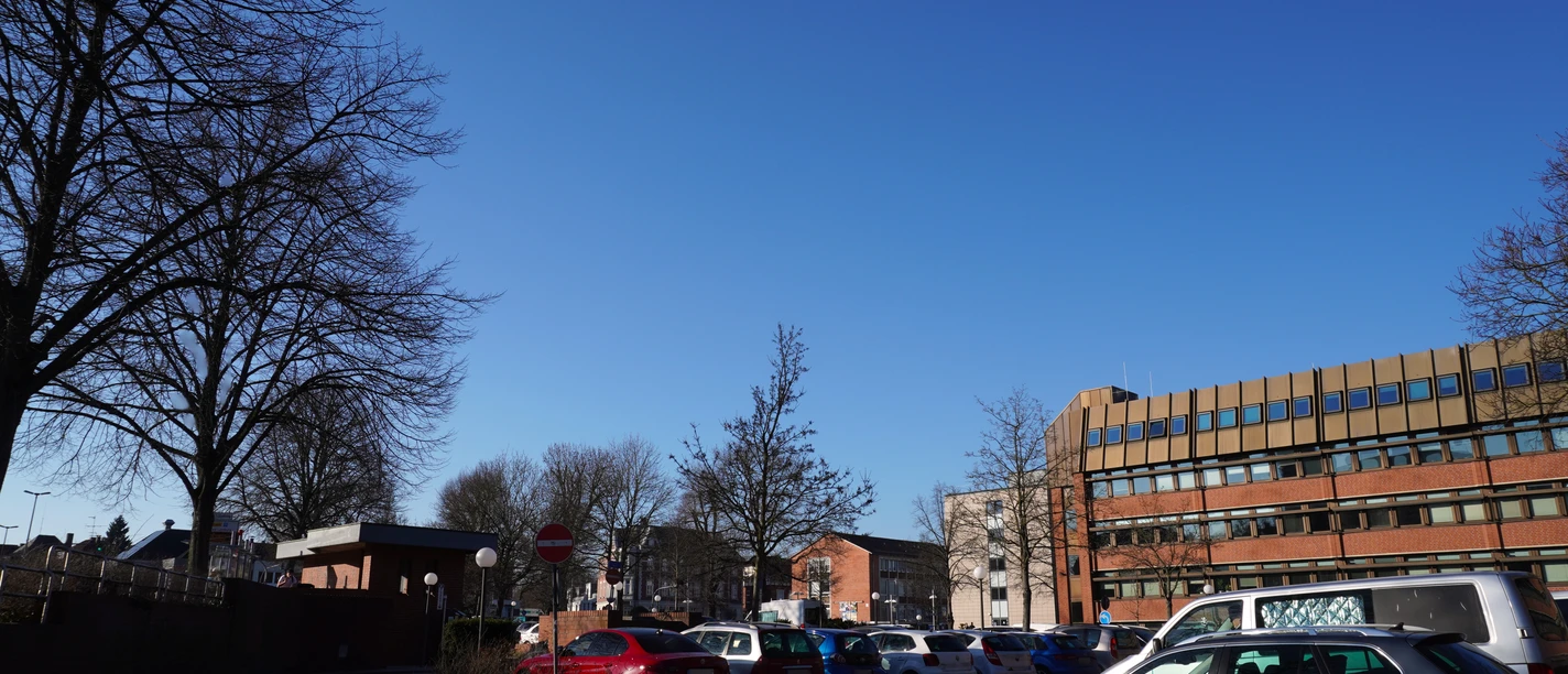 Geparkte Autos auf einem sonnigen Parkplatz vor einem mehrstöckigen Bürogebäude mit Bäumen.