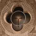 romanischer-kreuzweg.png Steinernes Vierpassfenster mit beleuchtetem Holzkreuz im Inneren eines Kirchenraums