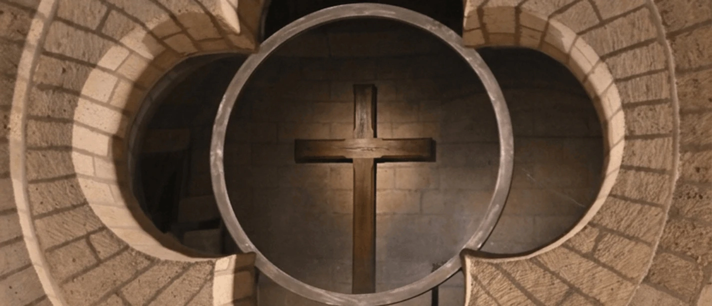 romanischer-kreuzweg.png Steinernes Vierpassfenster mit beleuchtetem Holzkreuz im Inneren eines Kirchenraums