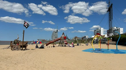 Spielplatz Weser-Strandbad Drachenfestival IMG_3401_ (c) Tanja Mehl_Erlebnis Bremerhaven_WEB.jpg Sonniger Strandspielplatz im Sand mit Holz-Piratenschiff, Rutsche und bunten Wasserspielen.