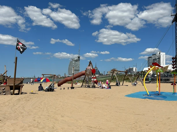 Spielplatz Weser-Strandbad Drachenfestival IMG_3401_ (c) Tanja Mehl_Erlebnis Bremerhaven_WEB.jpg Sonniger Strandspielplatz im Sand mit Holz-Piratenschiff, Rutsche und bunten Wasserspielen.