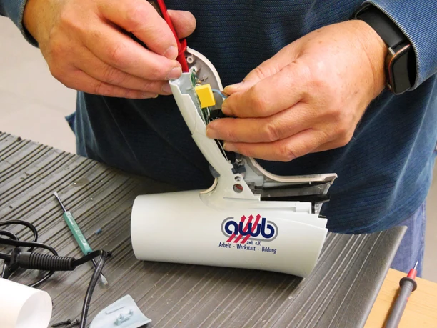 Eine Person arbeitet an einem technischen Gerät und verwendet dabei Werkzeuge. Auf dem Tisch sind weitere Werkzeuge und Teile sichtbar.
