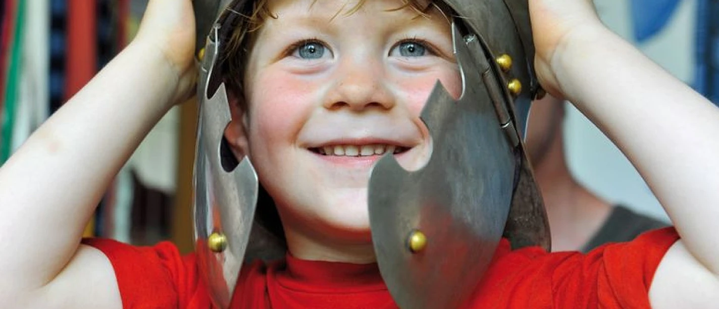 Familienführung in der Ausstellung zur Varusschlacht Ein lachender Junge trägt einen antiken Heldenhelm und blickt fröhlich in die Kamera.