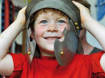 Familienführung in der Ausstellung zur Varusschlacht Ein lachender Junge trägt einen antiken Heldenhelm und blickt fröhlich in die Kamera.