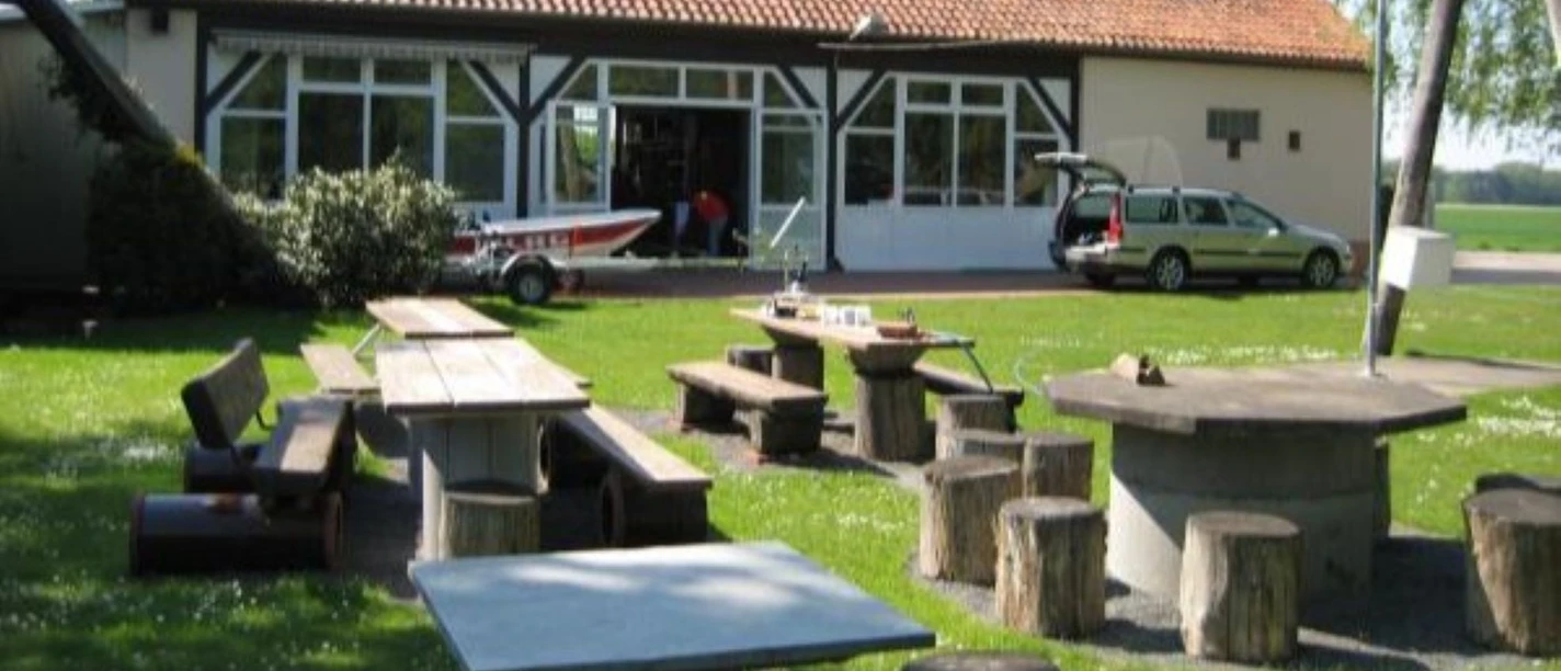 DLRG-Jugendbildungsstaette-Schirrhof_HD_16zu9.jpg DLRG-Jugendbildungsstätte mit Wiese, Steintischen, Holzhockern und einem Boot auf dem Hof.