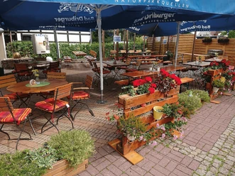 Biergarten in Kunkel´s Eck Biergarten mit Holztischen, roten Kissen, großen Sonnenschirmen und Blumenbeeten.