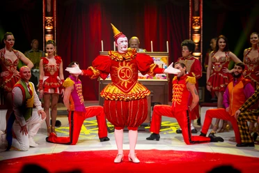 Opening, Circus Theater Roncalli Clown in red party costume stands in the spotlight, surrounded by artists