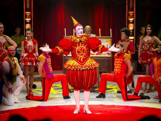 Opening, Circus Theater Roncalli Clown in red party costume stands in the spotlight, surrounded by artists