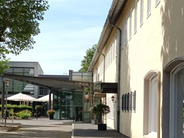 HotelamSee_HD_16zu9.jpg Eingangsbereich des Hotels am See in Salzgitter mit gelber Fassade, Vordach und alten Bäumen.