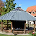 Runder Holz-Gradierpavillon mit Metalldach in einer Grünanlage vor einem Fachwerkhaus.