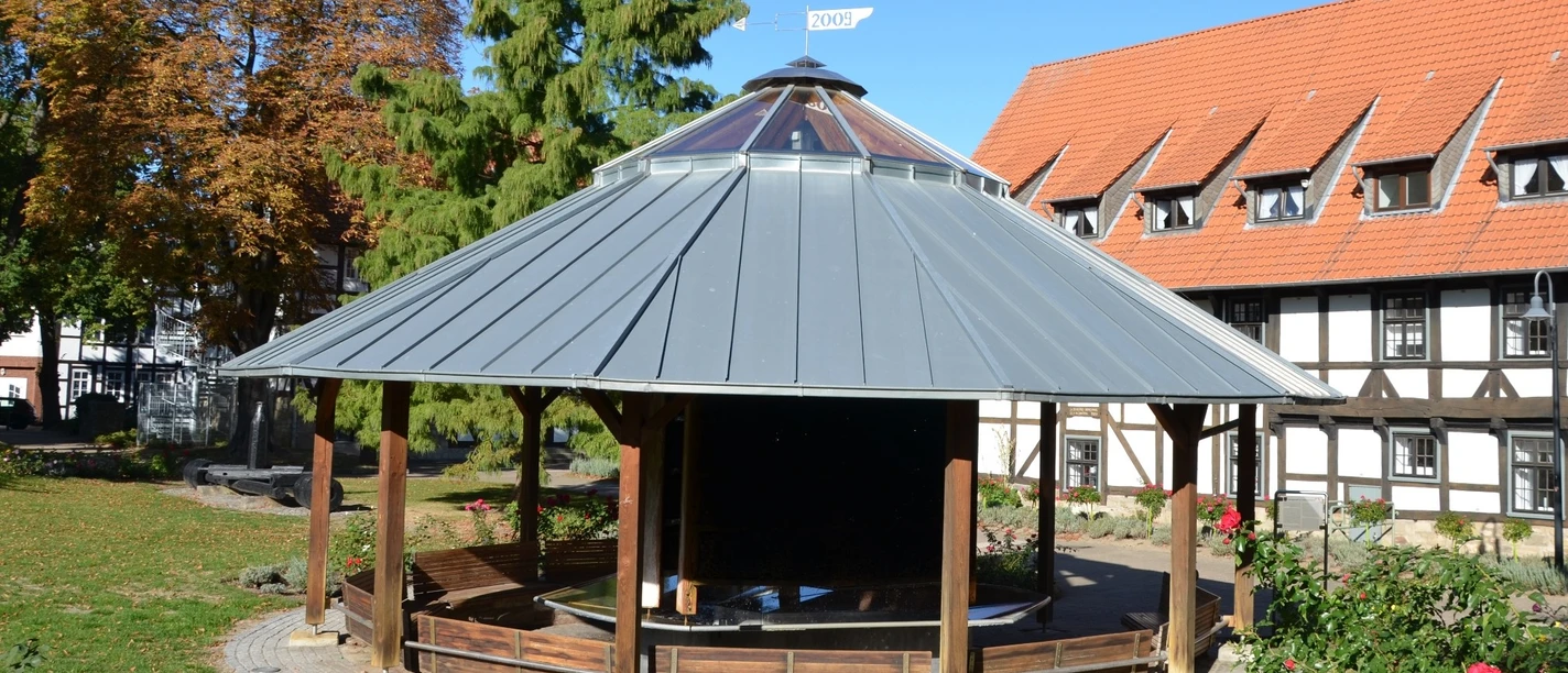 Gradierpavillon_Salzgitter_HD_16zu9.JPG Runder Holz-Gradierpavillon mit Metalldach in einer Grünanlage vor einem Fachwerkhaus.