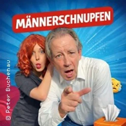 Ein Mann hustet theatralisch nach vorn, während eine Frau hinter ihm überrascht reagiert.