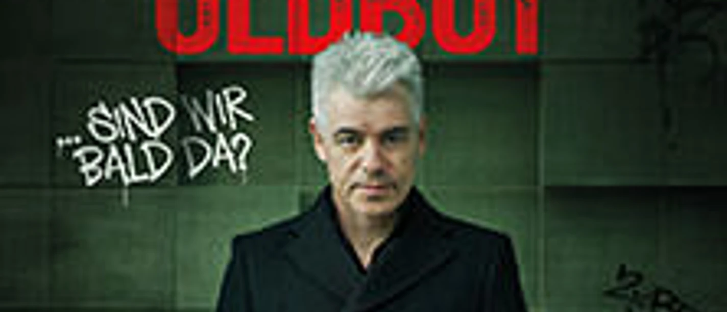 Ein Mann mit grauem Haar vor dunkler Wand und dem Schriftzug Oldboy sowie Sind wir bald da