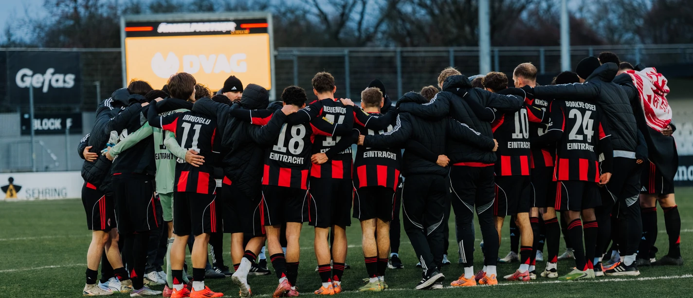 Football team in huddle on pitch, scoreboard behind