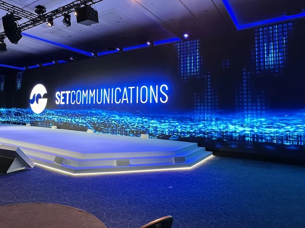 SETCOMMUNICATIONS GmbH - Modern event stage Modern event stage with a large LED screen showing the SETCOMMUNICATIONS logo and abstract blue light effects.