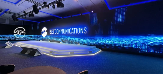 SETCOMMUNICATIONS GmbH - Moderne Eventbühne Moderne Eventbühne mit einer großen LED-Leinwand, die das Logo von SETCOMMUNICATIONS und abstrakte blaue Lichteffekte zeigt.