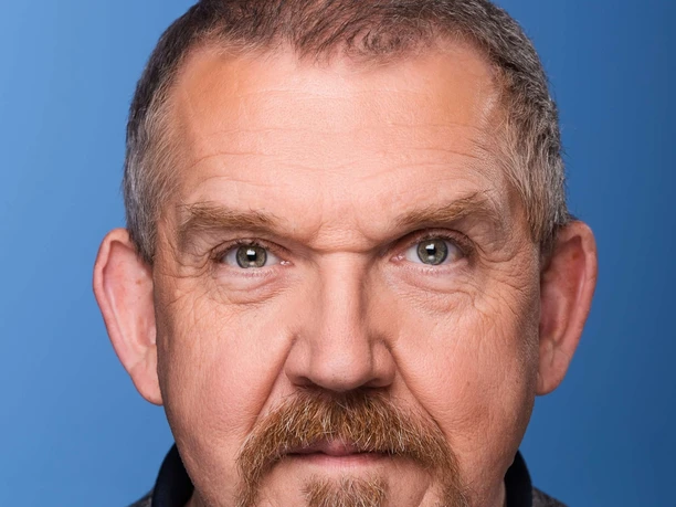 Dietmar Bär Porträt eines älteren Mannes mit kurzem Haar und Bart vor blauem Hintergrund.