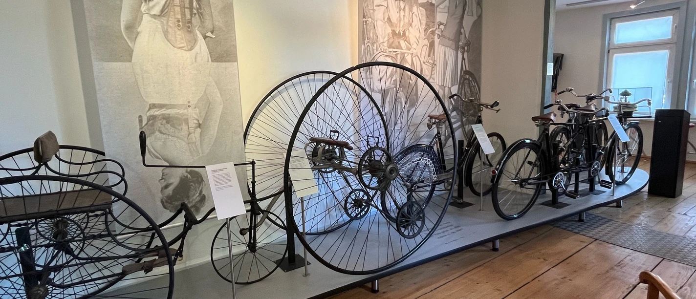 Radhaus 19.jpg Historische Fahrräder, darunter ein Hochrad, ausgestellt im Museum vor großformatigen Wandbildern.