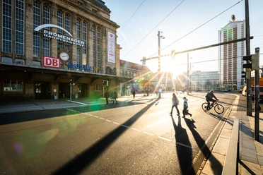 Promenaden Hauptbahnhof Leipzig - Nakupování v Lipsku Slunce vychází mezi lipským nádražím Promenaden Hauptbahnhof a věžákem Wintergarten, když kolemjdoucí přecházejí náměstí Willy-Brandt-Platz.