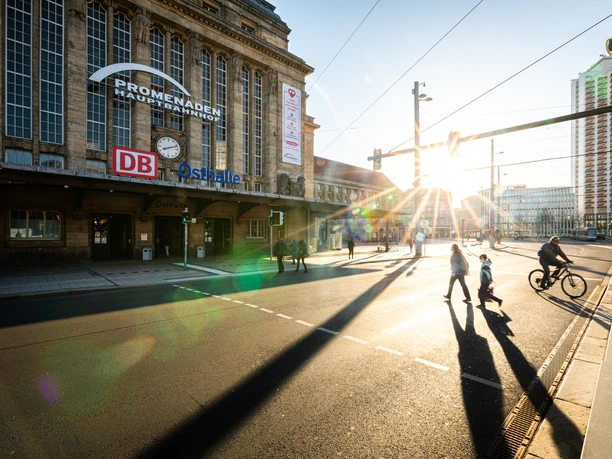 Promenaden Hauptbahnhof Leipzig - Shopping in Leipzig Die Sonne steigt zwischen dem Promenaden Hauptbahnhof Leipzig und dem Wintergartenhochhaus auf, während Passanten den Willy-Brandt-Platz überqueren.
