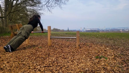 Trimmpfad - Liegestütz-Station.jpg Frau macht Liegestütze an einer Holzstation im Wald am Klieversberg mit Blick auf Wolfsburg.