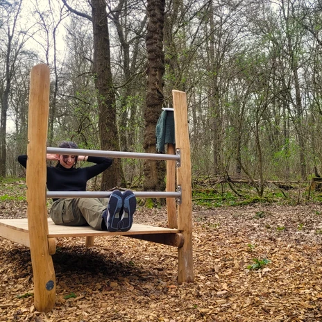 Trimmpfad - Sit-up-Station.jpg Frau macht Sit-ups an einer hölzernen Fitnessstation im Wald.