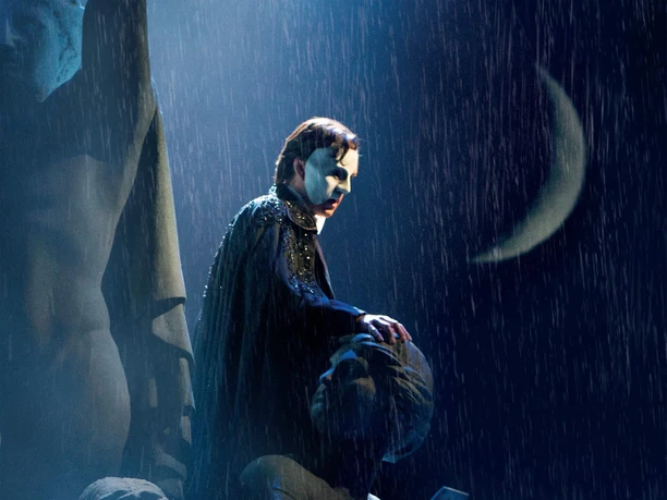 Das Phantom der Oper Vermummte Bühnenfigur mit Halbmaske steht im Regen zwischen dunklen Statuen vor Mondsichel.