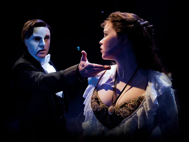 The Phantom of the Opera Masked man holds out his hand to a young woman in the dark stage light