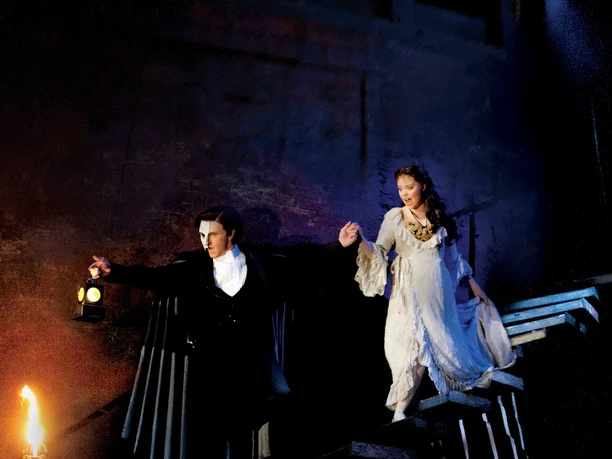 The Phantom of the Opera Two figures descend a staircase in a dark scene, surrounded by fog and torchlight