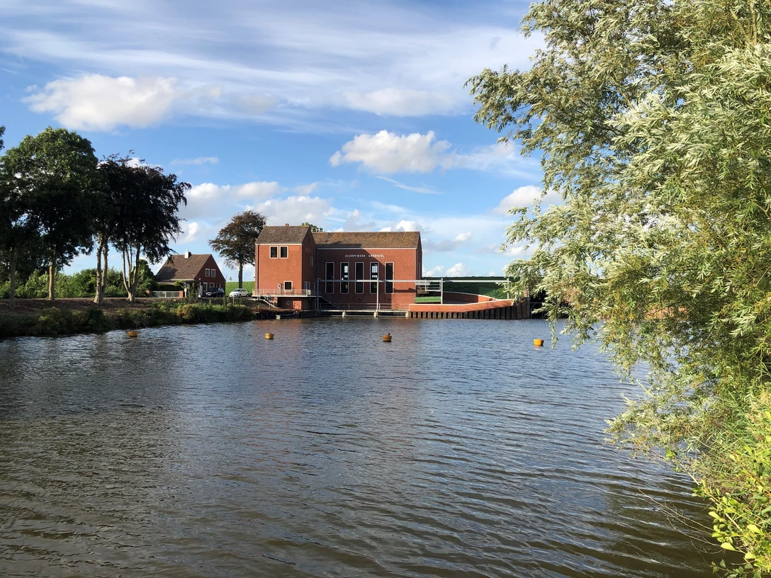 Schöpfwerk Greetsiel Backsteinbau des Schöpfwerks Greetsiel am Wasser, umgeben von Bäumen unter blauem Himmel.