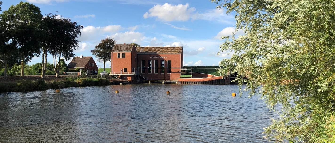 Schöpfwerk Greetsiel Backsteinbau des Schöpfwerks Greetsiel am Wasser, umgeben von Bäumen unter blauem Himmel.