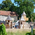 Ritterburgspielplatz1_HD_16zu9.JPG Großer hölzerner Ritterburgspielplatz mit Türmen und Rutsche vor historischen Steinmauern.