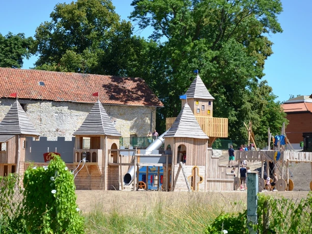 Ritterburgspielplatz1_HD_16zu9.JPG Großer hölzerner Ritterburgspielplatz mit Türmen und Rutsche vor historischen Steinmauern.