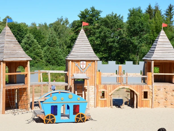 Ritterburgspielplatz2_HD_16zu9.JPG Großer Holzspielplatz als Ritterburg mit drei Türmen und einer blauen Spiel-Kutsche im Sand.