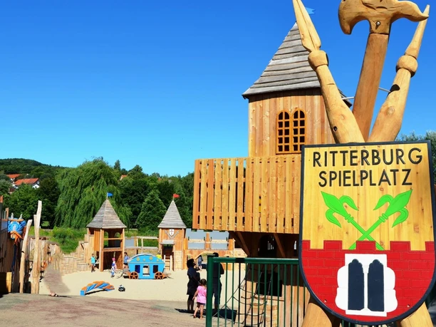 Ritterburgspielplatz3_HD_16zu9.jpg Hölzerner Spielplatz als Ritterburg mit Türmen und Wappenschild im Vordergrund.