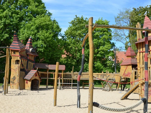 Roxy-Helco-Spielplatz_HD_16zu9.JPG Großer Abenteuerspielplatz aus Holz in Burgoptik mit roten Dächern, Sandboden und grünen Bäumen.