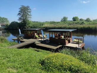 Zwei überdachte Holzflöße liegen an einem Steg am ruhigen Flussufer in sonniger Naturlandschaft.