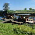 NoBoat Flöße Otterndorf Zwei überdachte Holzflöße liegen an einem Steg am ruhigen Flussufer in sonniger Naturlandschaft.