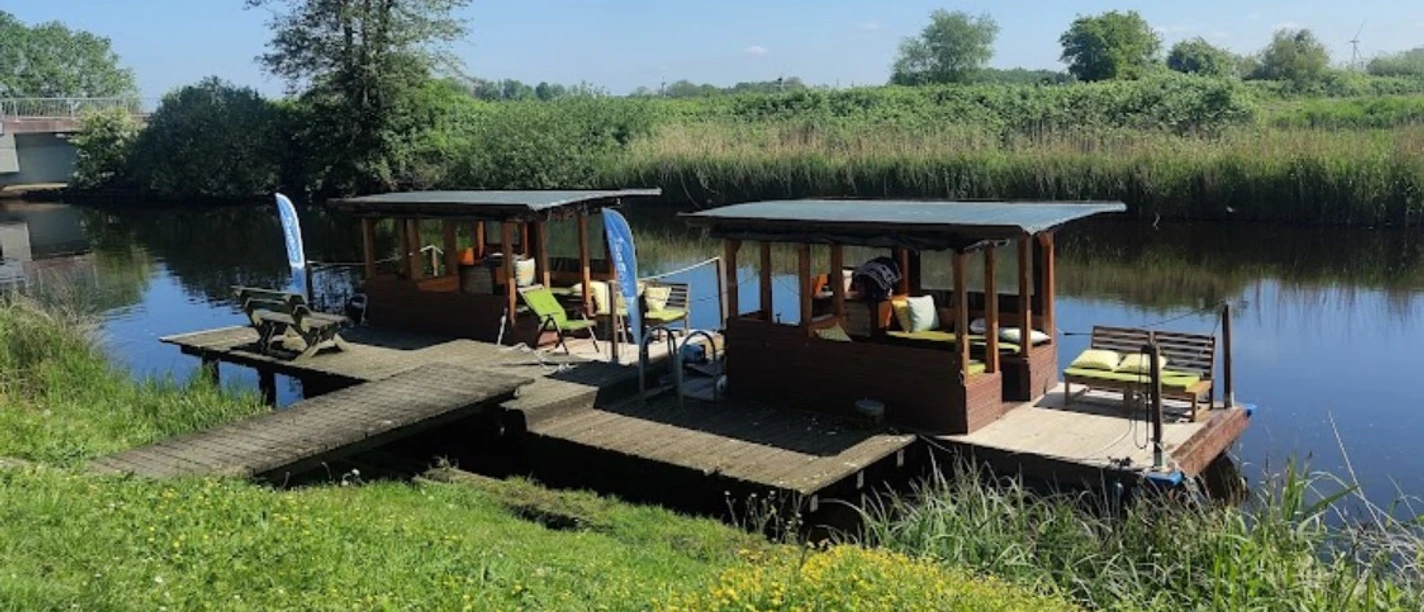 NoBoat Flöße Otterndorf Zwei überdachte Holzflöße liegen an einem Steg am ruhigen Flussufer in sonniger Naturlandschaft.