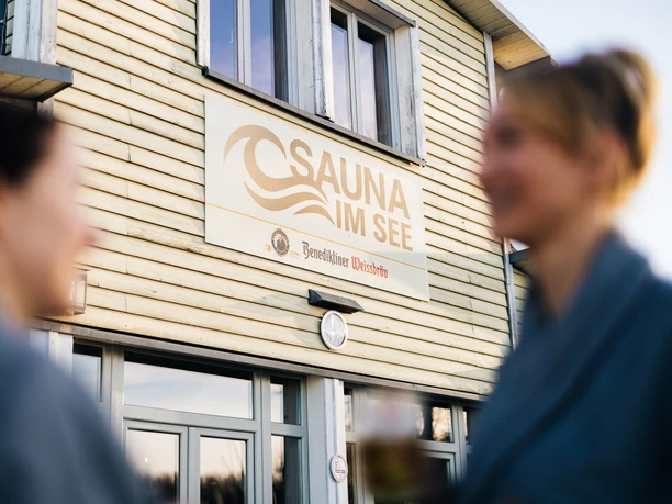 Sauna im See - Wellness in Leipzig und Region Zwei Frauen in Bademänteln unterhalten sich im Außenbereich auf der Terrasse der Sauna im See, deren Schriftzug groß auf der Fassade hängt.