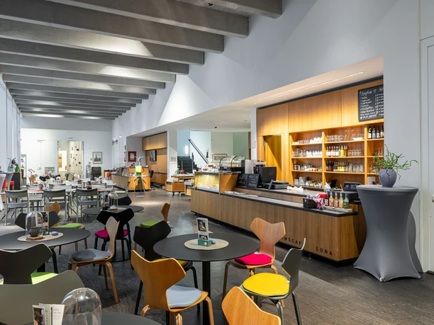 Heller Innenraum des Museumscafés LUNA mit modernem Tresen und bunten Stühlen an runden Tischen.