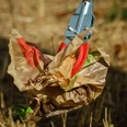 Müll aufsammeln Eine Greifzange hebt zerknülltes, farbig bedrucktes Papier von trockenem Grasboden auf.