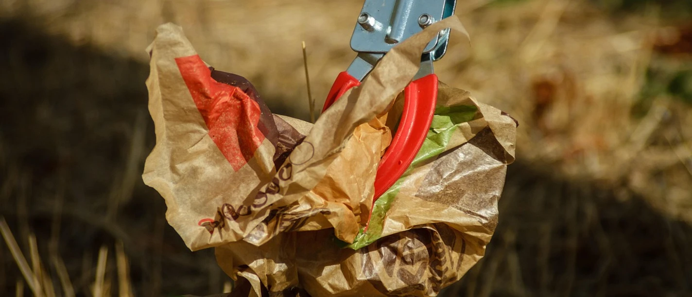 Müll aufsammeln Eine Greifzange hebt zerknülltes, farbig bedrucktes Papier von trockenem Grasboden auf.