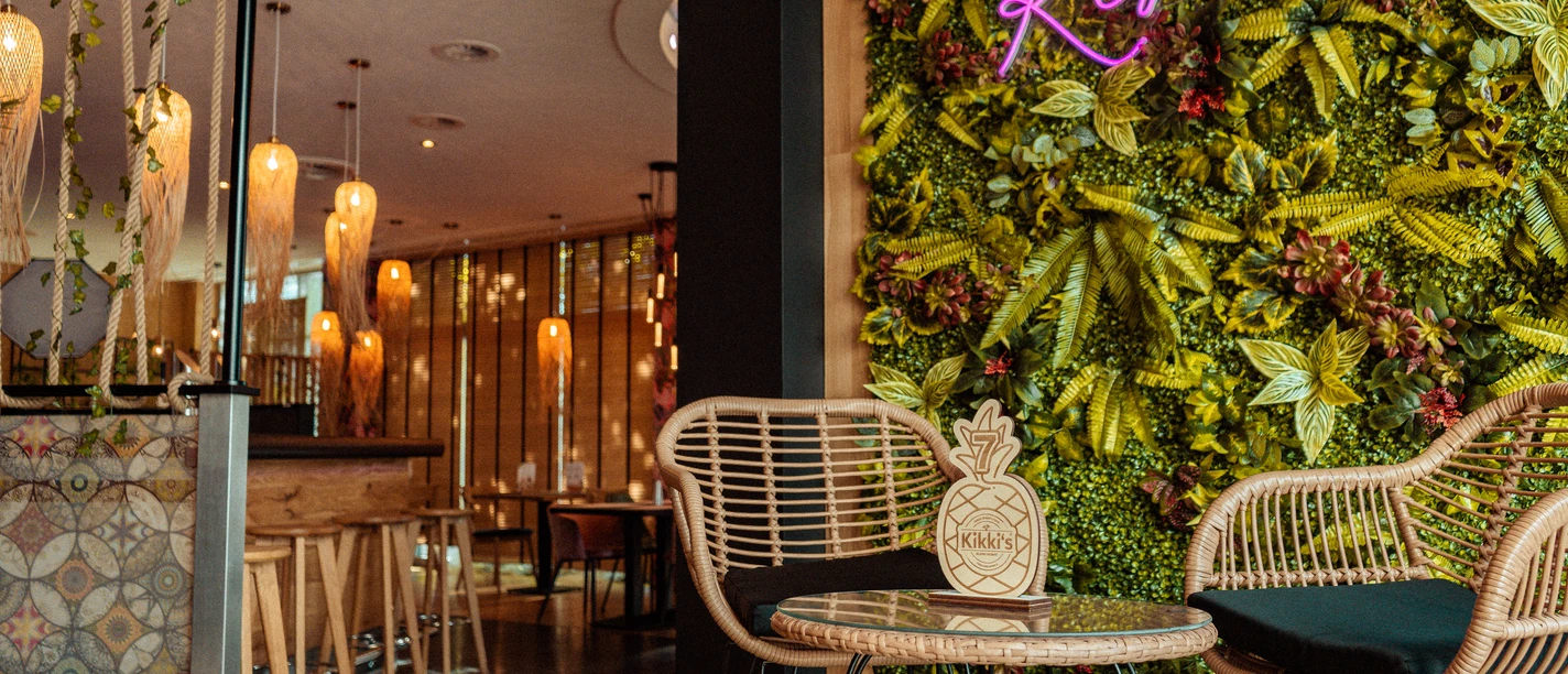 Kikkis_Damme Rattanstühle am Glastisch vor einer grünen Pflanzenwand mit pinkem Kikki's Neonlicht.