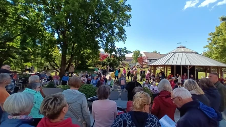2025_Buergersingen_Rosengarten.jpg Eine Menschenmenge singt mit Textblättern im sonnigen Park vor einem Holzpavillon.