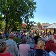 2025_Buergersingen_Rosengarten.jpg Eine Menschenmenge singt mit Textblättern im sonnigen Park vor einem Holzpavillon.