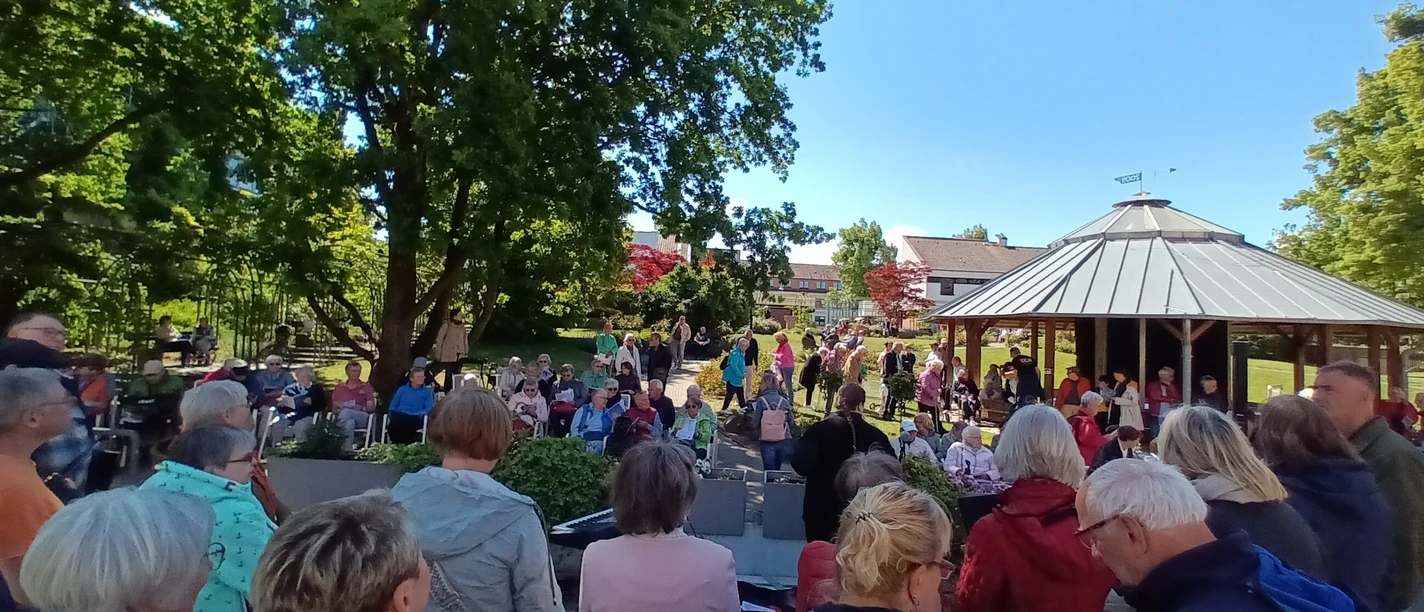 2025_Buergersingen_Rosengarten.jpg Eine Menschenmenge singt mit Textblättern im sonnigen Park vor einem Holzpavillon.