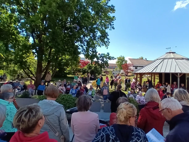 2025_Buergersingen_Rosengarten.jpg Eine Menschenmenge singt mit Textblättern im sonnigen Park vor einem Holzpavillon.