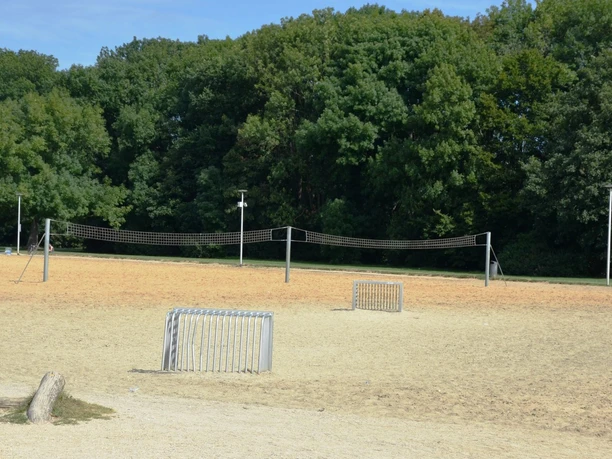 Beachvolleyballfeld_Salzgittersee_HD_16zu9.JPG Weites Sandfeld am Salzgittersee mit Beachvolleyballnetz und zwei Toren vor grünen Bäumen.
