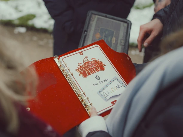 Epic Escape Roter Ordner mit Aufgabenseite zu Mission Weihnachtsfeier, umringt von Händen im Freien.